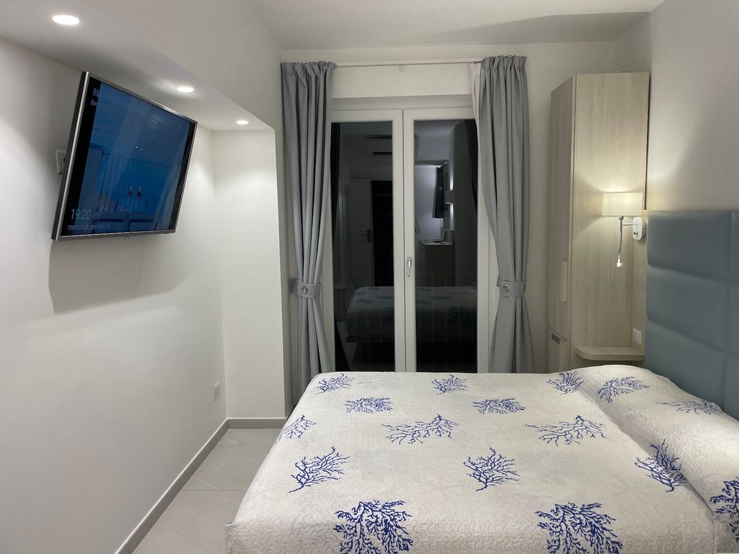 A modern hotel bedroom featuring a queen bed with blue patterned bedding, wall-mounted television, mirrored wardrobe, and access to a balcony with gray curtains.