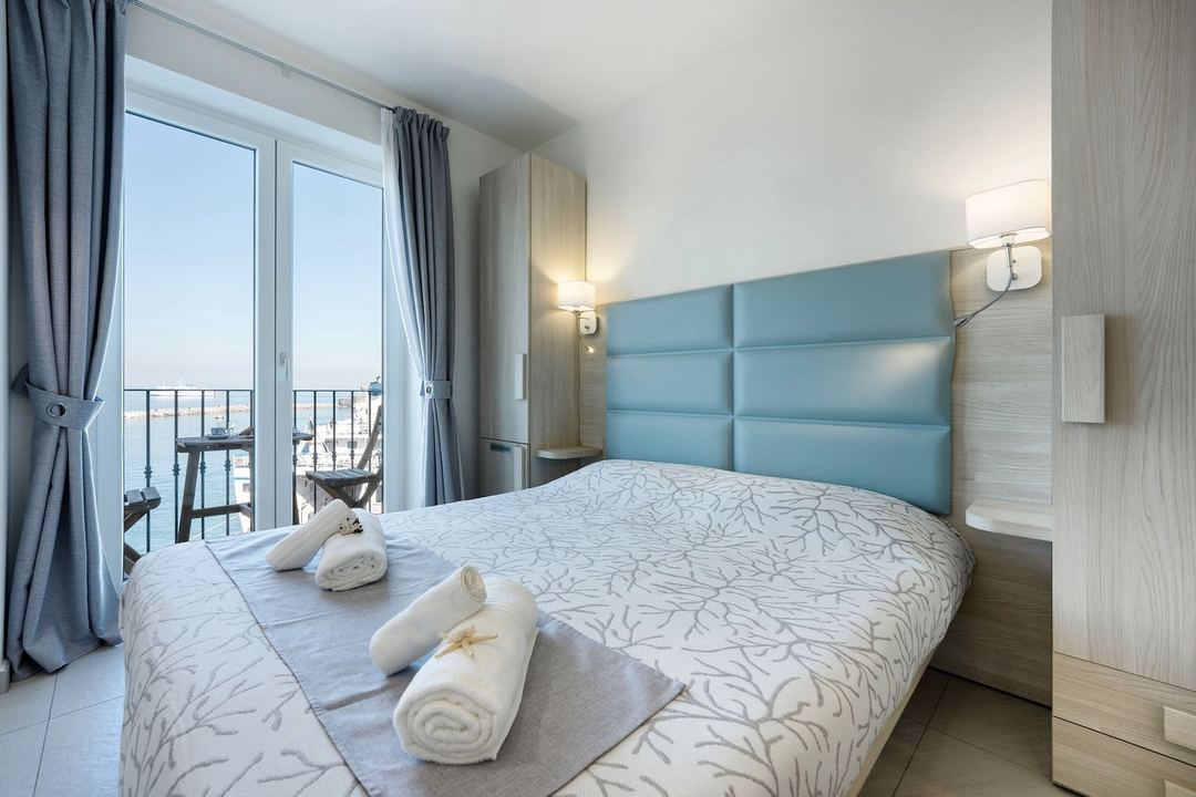 A modern hotel bedroom with a blue upholstered headboard, sea view balcony, and rolled towels arranged on the bed.