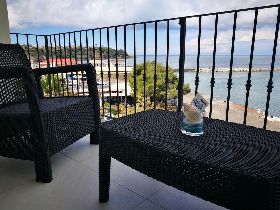 Scenic balcony view of the sea and coastline from a hotel terrace furnished with black wicker seating.