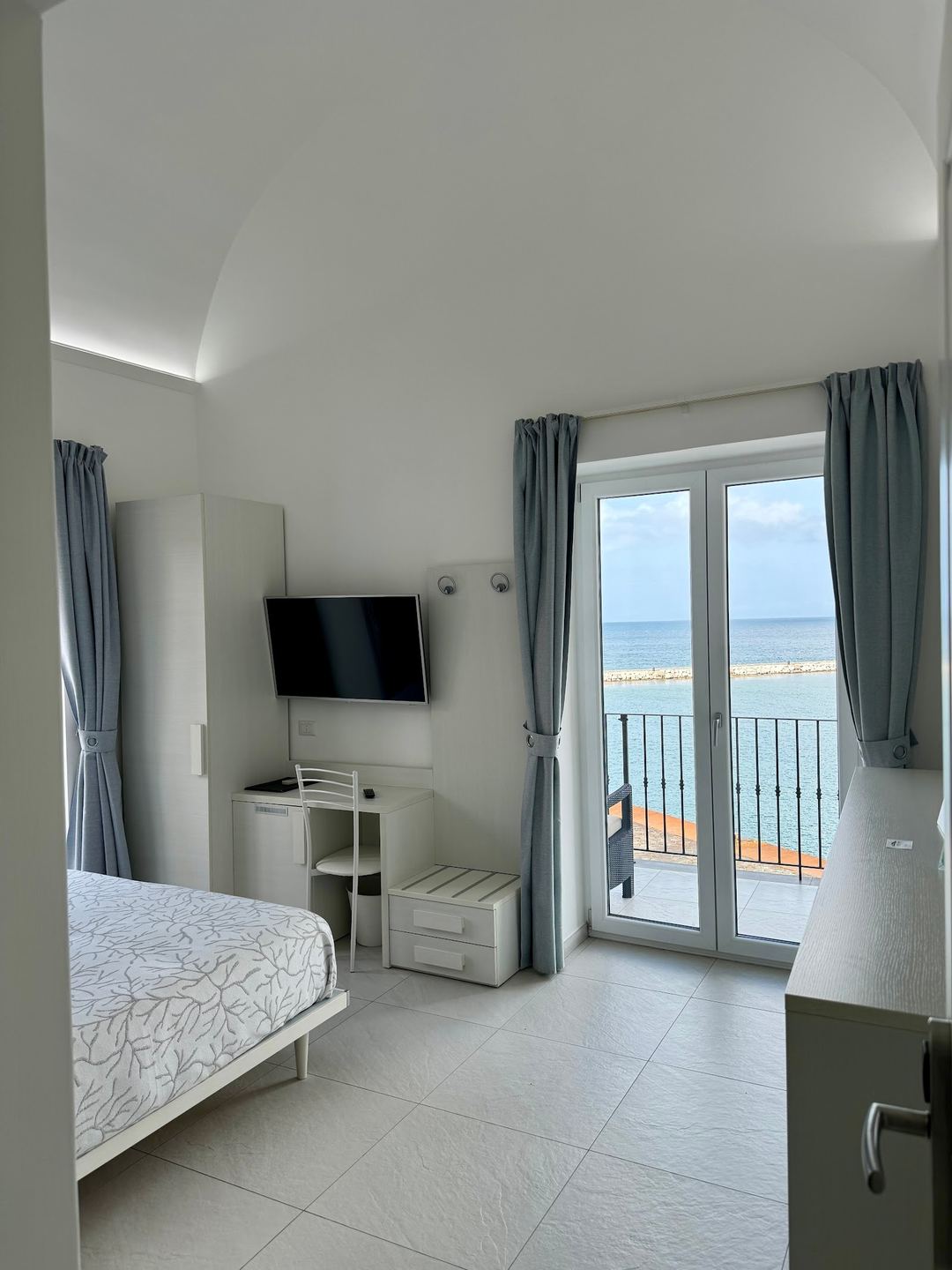 A modern beachfront hotel room with a bed, wall-mounted TV, and sea view through glass doors opening to a balcony.