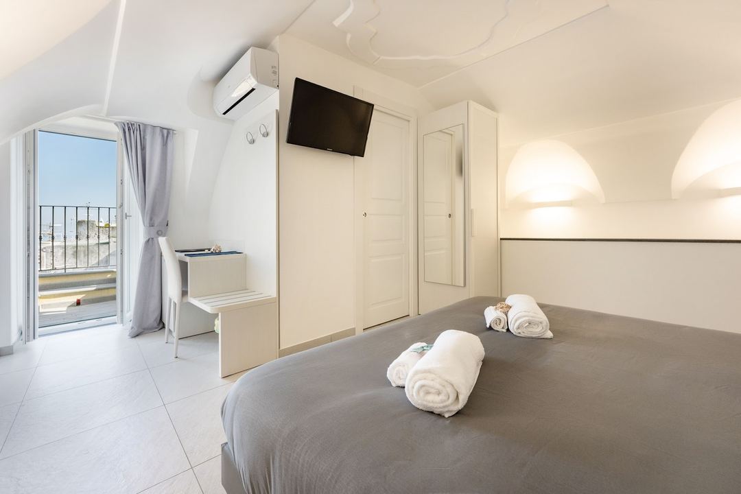 A bright, minimalist hotel bedroom with a gray bed, white rolled towels, flat-screen TV, and access to a sea-view balcony.