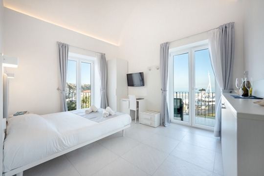 A bright, modern bedroom with a white bed, dual glass doors opening to a sea-view balcony, and minimalist décor.